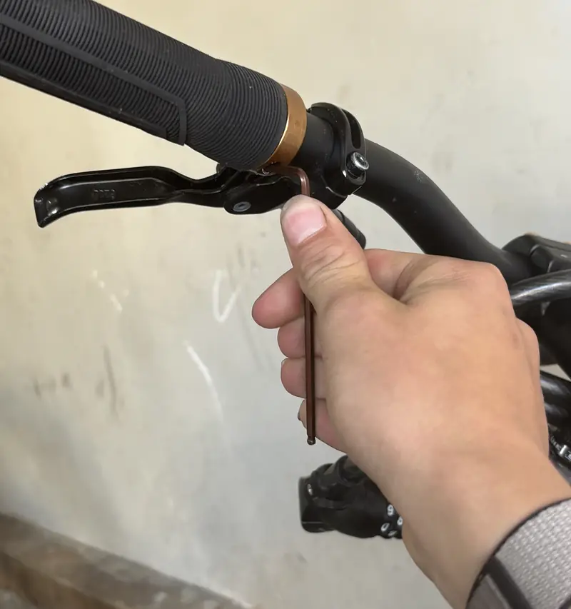 A hex key adjusting a hydraulic brake lever on a black handlebar.